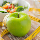 A bowl of salad, slightly blurred, sits behind a green apple with a yellow tape measure loosely wrapped around it.
