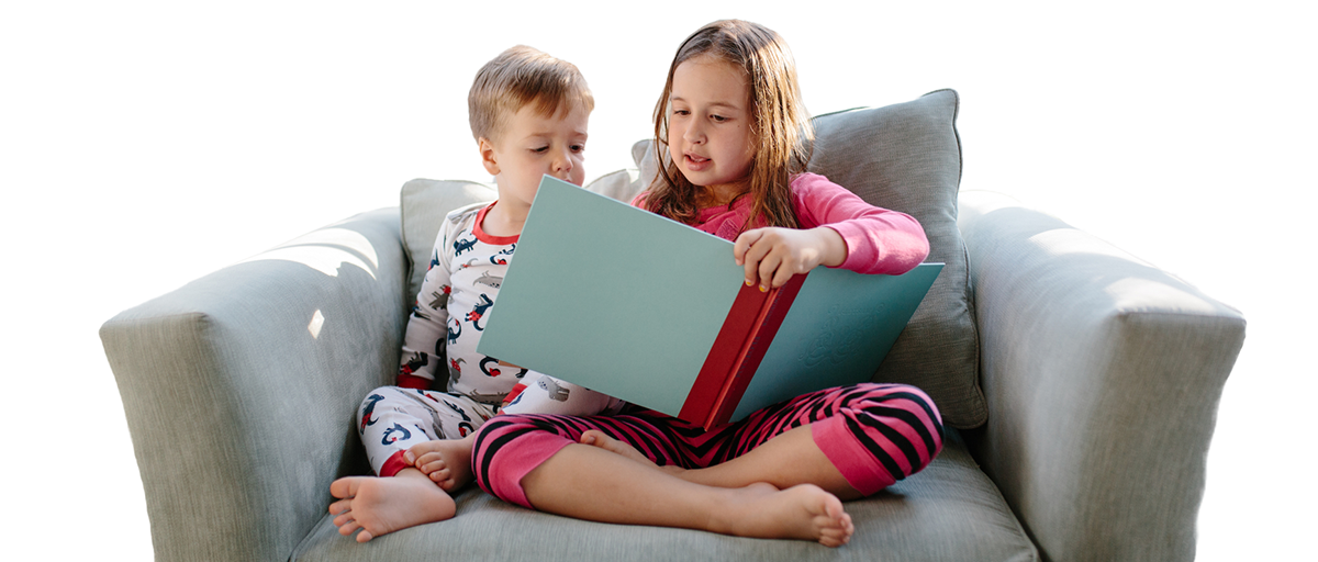 Two children, one older in age, share a couch while reading a book together.